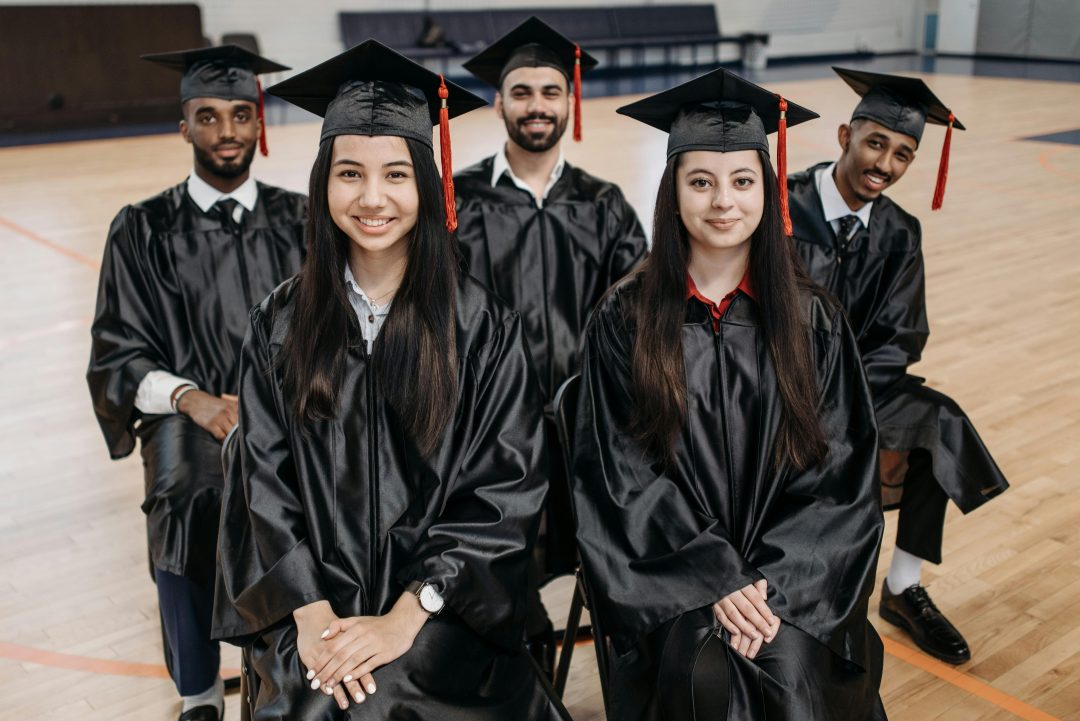 A diverse group of young students wearing convocation caps and gowns, smiling at the camera.