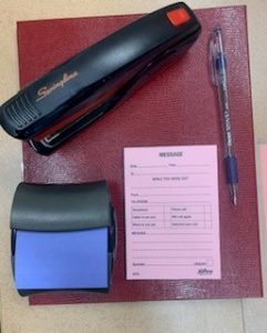 A stapler, a blue sticky note in a dispenser, a pink message pad and a ball point pen on a maroon notebook.