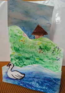 Folded paper has been cut and coloured to create a 3-D illustration. The foreground has a swan swimming in water. The middle ground is a green, grassy riverbank with yellow, pink and purple flowers. The is a brown hut at the top of the riverbank. The background is a blue sky with several shades of blue and 3 white clouds.