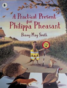 The illustrated cover of a children's book called "A Practical Present for Philippa Pheasant" by Briony May Smith is shown. The picture features a pheasant in a yellow safety vest holding a stop sign. A mouse is smiling up at the pheasant. In front of them is a crosswalk with a hedgehog, weasel, and a grouse crossing the road from left to right. There are hedges along the side of the road and a house with a thatched roof in the background. The lower trunk of a large tree is in the upper right corner and it has golden leaves on a branch that extend to the top middle of the cover, above the text. A few leaves are midair on the upper left side.