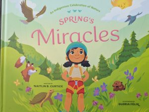 The cover of the picture book "Spring's Miracles" is shown. There is small green text at the centre of the top that reads "An Indigenous Celebration of Nature." Under that is larger text with the title in pink. In the centre is a child dressed in shorts and a t-shirt with a helmet and a climbing harness. She is a field of grass, flowers and animls including a fox, turtle and groundhog. There are birds in the sky as well as a smiling sun and clouds. There are trees and mountains in the background. Written by Kaitlin B. Curtice is the black text over the turtle on the left and Illustrated by Gloria Felix is in the lower right corner.