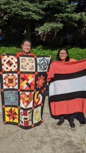 A photograph of two First Nations women is shown. They are standing outside and holding up a handmade blanket and quilt that feature the colour orange. The quilt has 12 squares including a heart near the centre.