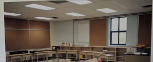 Classroom with bare walls and bulletin boards is shown in a photograph. There are a few student desks and a teacher desk but no resources.