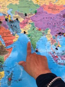 An index finger of a right hand pointing to Mumbai on the West Coast of India. The arm is in a black sleeve on which there is a silver bracelet.
