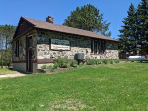 A small stone building with a sign saying "Haliburton County Library Stanhope Branch". Green grass in the foreground and a bright blue sky.