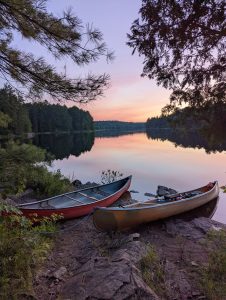 A photograph of a sunset reflecting on a tree-lined lake with two canoes resting on the shore in the foreground.